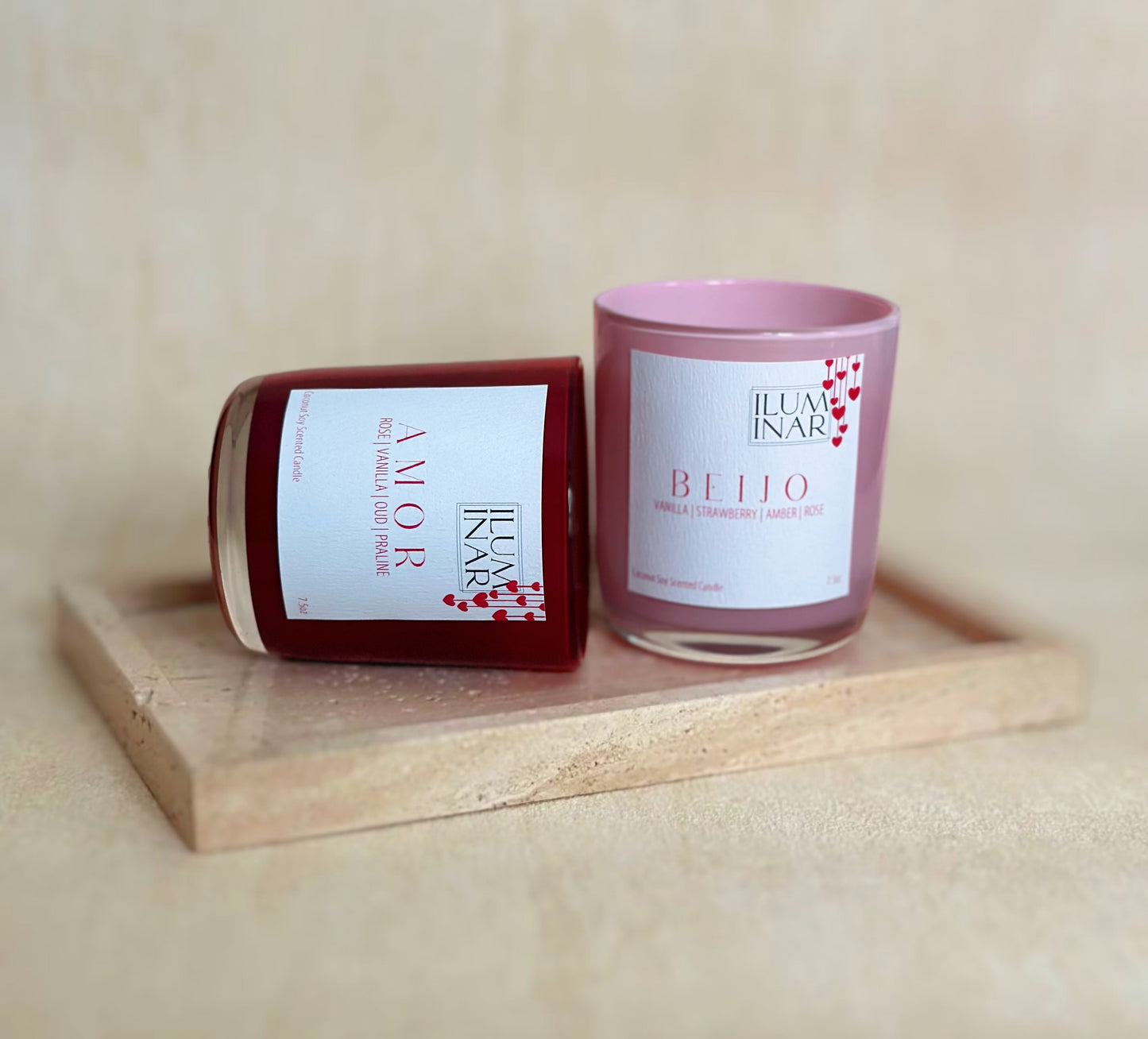 A beautifully styled product photo featuring Iluminar’s luxury scented candles, AMOR and BEIJO. The AMOR candle is housed in a deep red, glossy glass vessel, while BEIJO is in a soft pink vessel. Both candles are elegantly placed on a neutral-toned textured surface with a marble tray, creating a sophisticated and romantic ambiance. The labels showcase their scent notes: AMOR with rose, vanilla, oud, and praline; BEIJO with vanilla, strawberry, amber, and rose. The image is softly lit for a high-end, profes