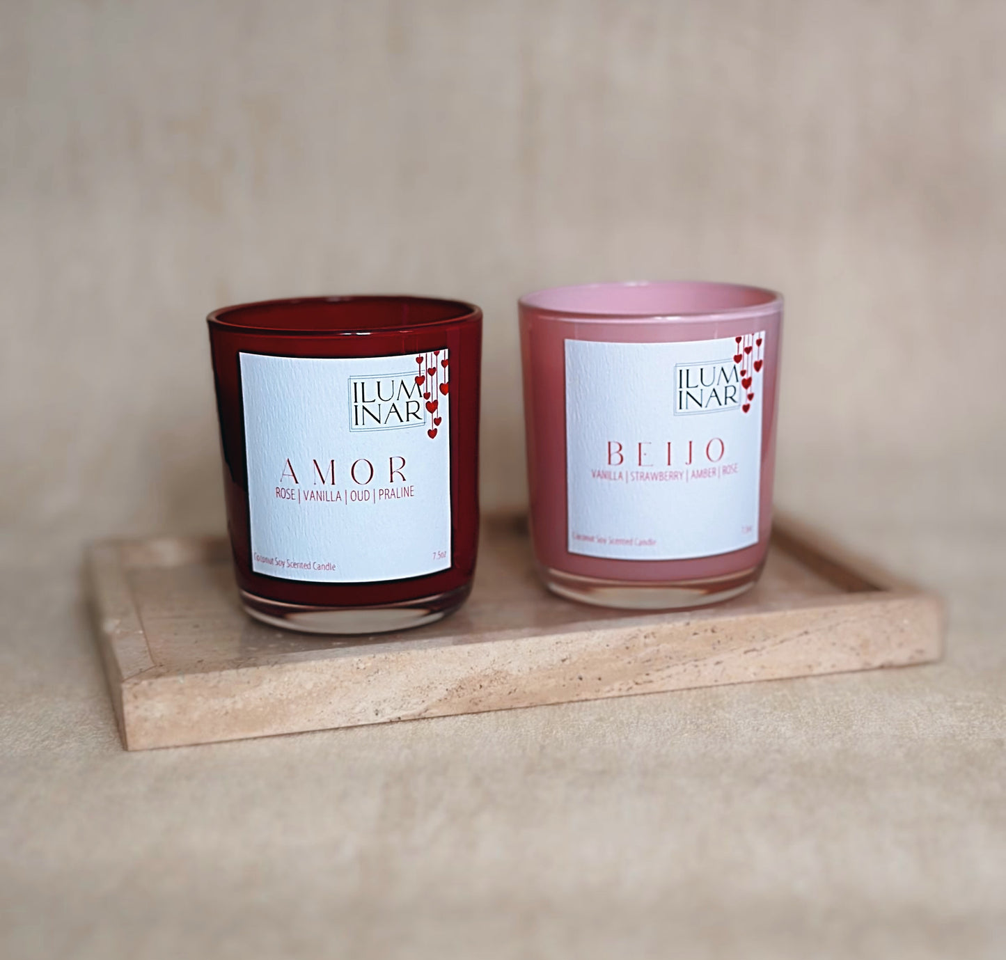A beautifully styled product photo featuring Iluminar’s luxury scented candles, AMOR and BEIJO. The AMOR candle is housed in a deep red, glossy glass vessel, while BEIJO is in a soft pink vessel. Both candles are elegantly placed on a neutral-toned textured surface with a marble tray, creating a sophisticated and romantic ambiance. The labels showcase their scent notes: AMOR with rose, vanilla, oud, and praline; BEIJO with vanilla, strawberry, amber, and rose. The image is softly lit for a high-end, profes