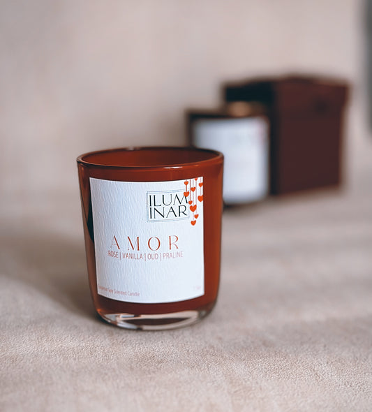A beautifully styled product photo featuring Iluminar’s luxury scented candles, AMOR and BEIJO. The AMOR candle is housed in a deep red, glossy glass vessel, while BEIJO is in a soft pink vessel. Both candles are elegantly placed on a neutral-toned textured surface with a marble tray, creating a sophisticated and romantic ambiance. The labels showcase their scent notes: AMOR with rose, vanilla, oud, and praline; BEIJO with vanilla, strawberry, amber, and rose. The image is softly lit for a high-end, profes