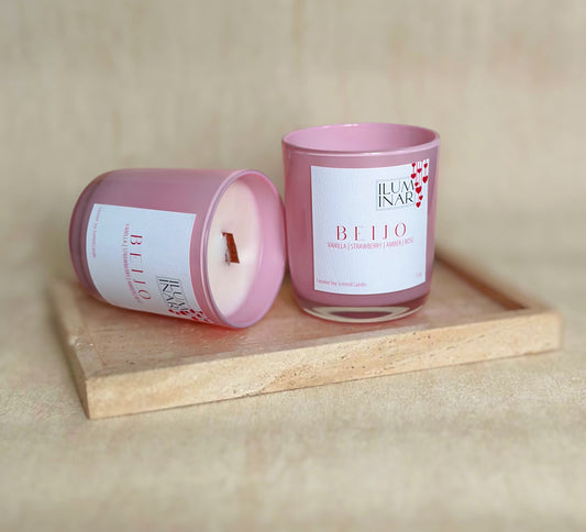 A beautifully styled product photo featuring Iluminar’s luxury scented candles, AMOR and BEIJO. The AMOR candle is housed in a deep red, glossy glass vessel, while BEIJO is in a soft pink vessel. Both candles are elegantly placed on a neutral-toned textured surface with a marble tray, creating a sophisticated and romantic ambiance. The labels showcase their scent notes: AMOR with rose, vanilla, oud, and praline; BEIJO with vanilla, strawberry, amber, and rose. The image is softly lit for a high-end, profes