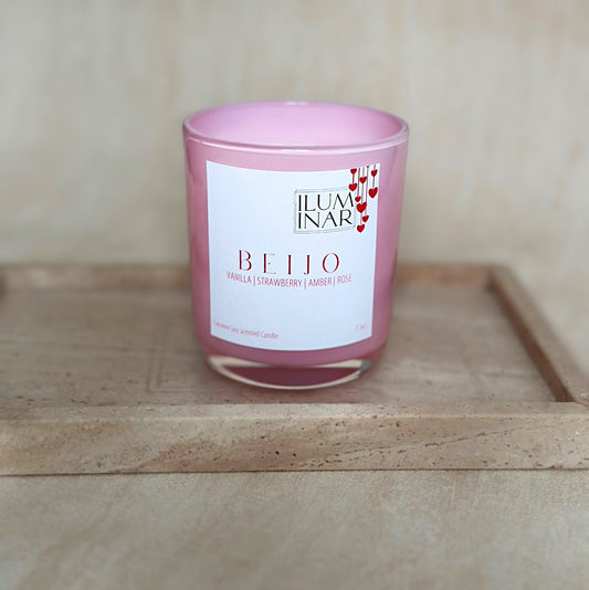 A beautifully styled product photo featuring Iluminar’s luxury scented candles, AMOR and BEIJO. The AMOR candle is housed in a deep red, glossy glass vessel, while BEIJO is in a soft pink vessel. Both candles are elegantly placed on a neutral-toned textured surface with a marble tray, creating a sophisticated and romantic ambiance. The labels showcase their scent notes: AMOR with rose, vanilla, oud, and praline; BEIJO with vanilla, strawberry, amber, and rose. The image is softly lit for a high-end, profes
