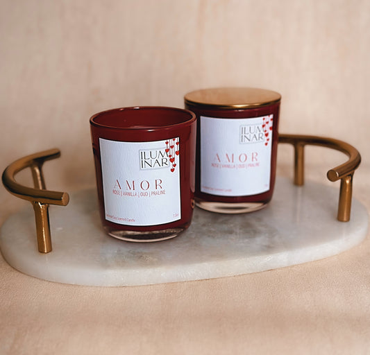 A beautifully styled product photo featuring Iluminar’s luxury scented candles, AMOR and BEIJO. The AMOR candle is housed in a deep red, glossy glass vessel, while BEIJO is in a soft pink vessel. Both candles are elegantly placed on a neutral-toned textured surface with a marble tray, creating a sophisticated and romantic ambiance. The labels showcase their scent notes: AMOR with rose, vanilla, oud, and praline; BEIJO with vanilla, strawberry, amber, and rose. The image is softly lit for a high-end, profes
